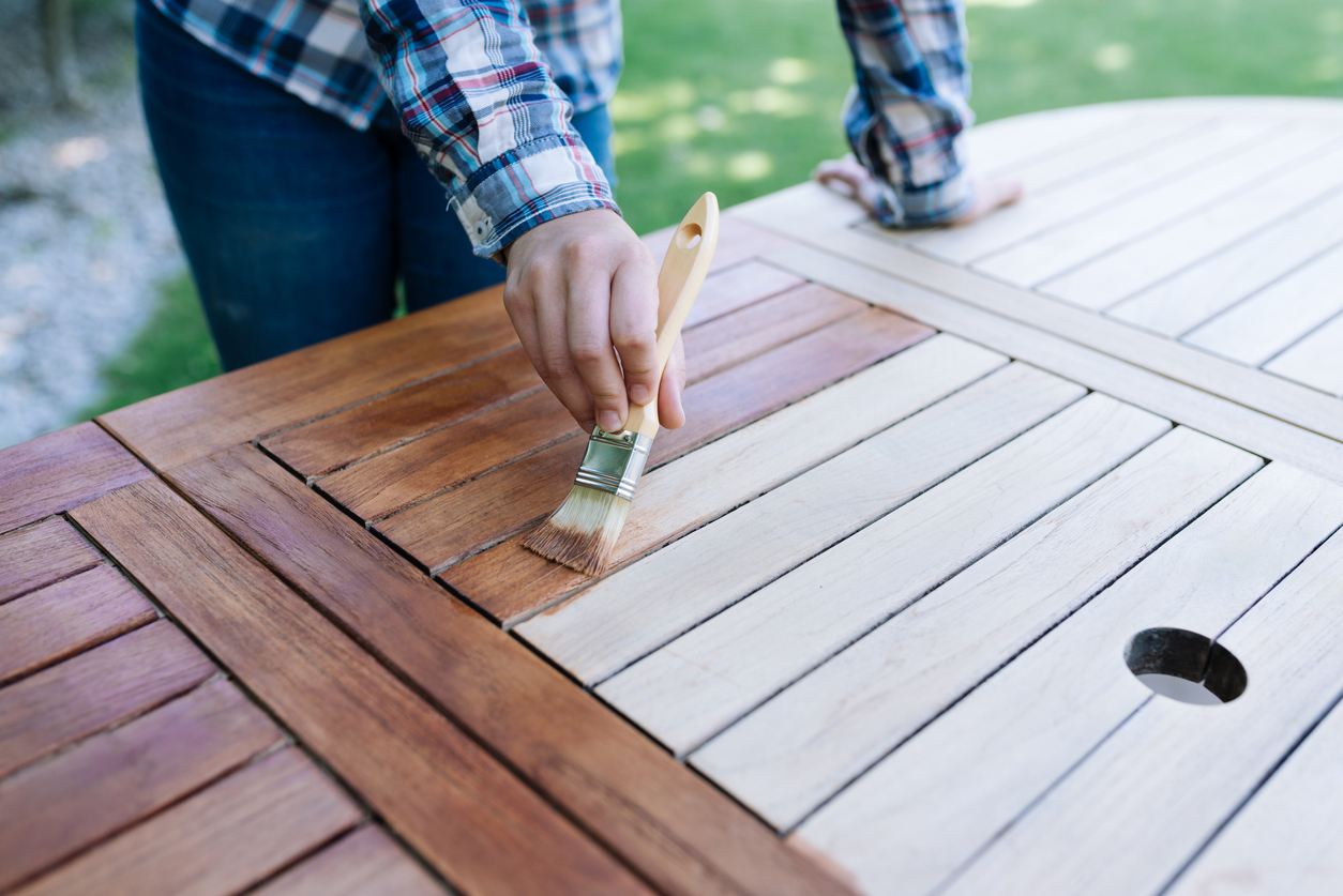 Person trägt Holzöl mit einem Pinsel auf einen Gartentisch aus Holz auf.