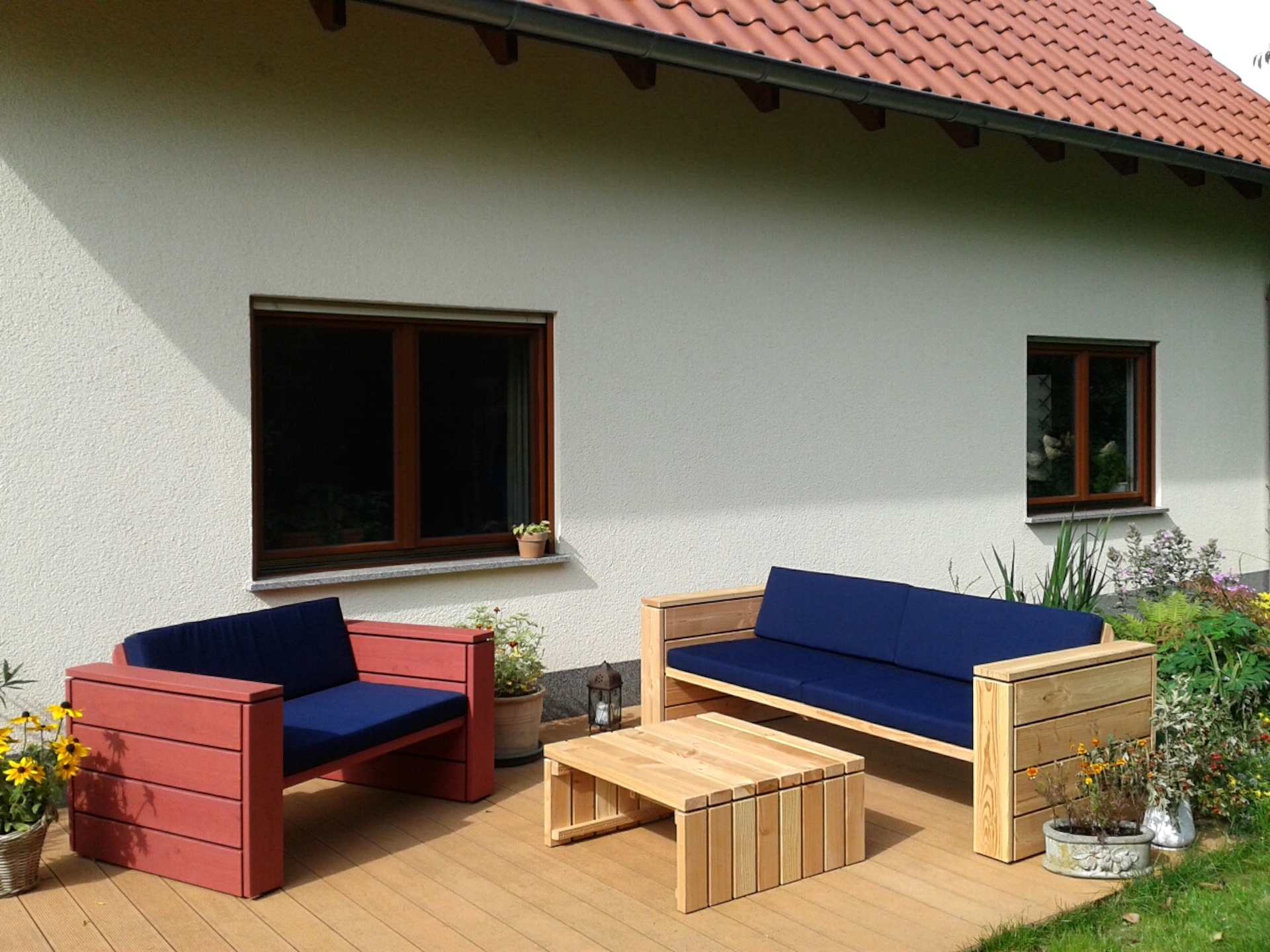 Bunte Loungemöbel: 2er Sofa in nordisch rot, 3er Sofa und Tisch natur geölt, Polster in Dunkelblau, auf Holzterrasse vor Putzfassade