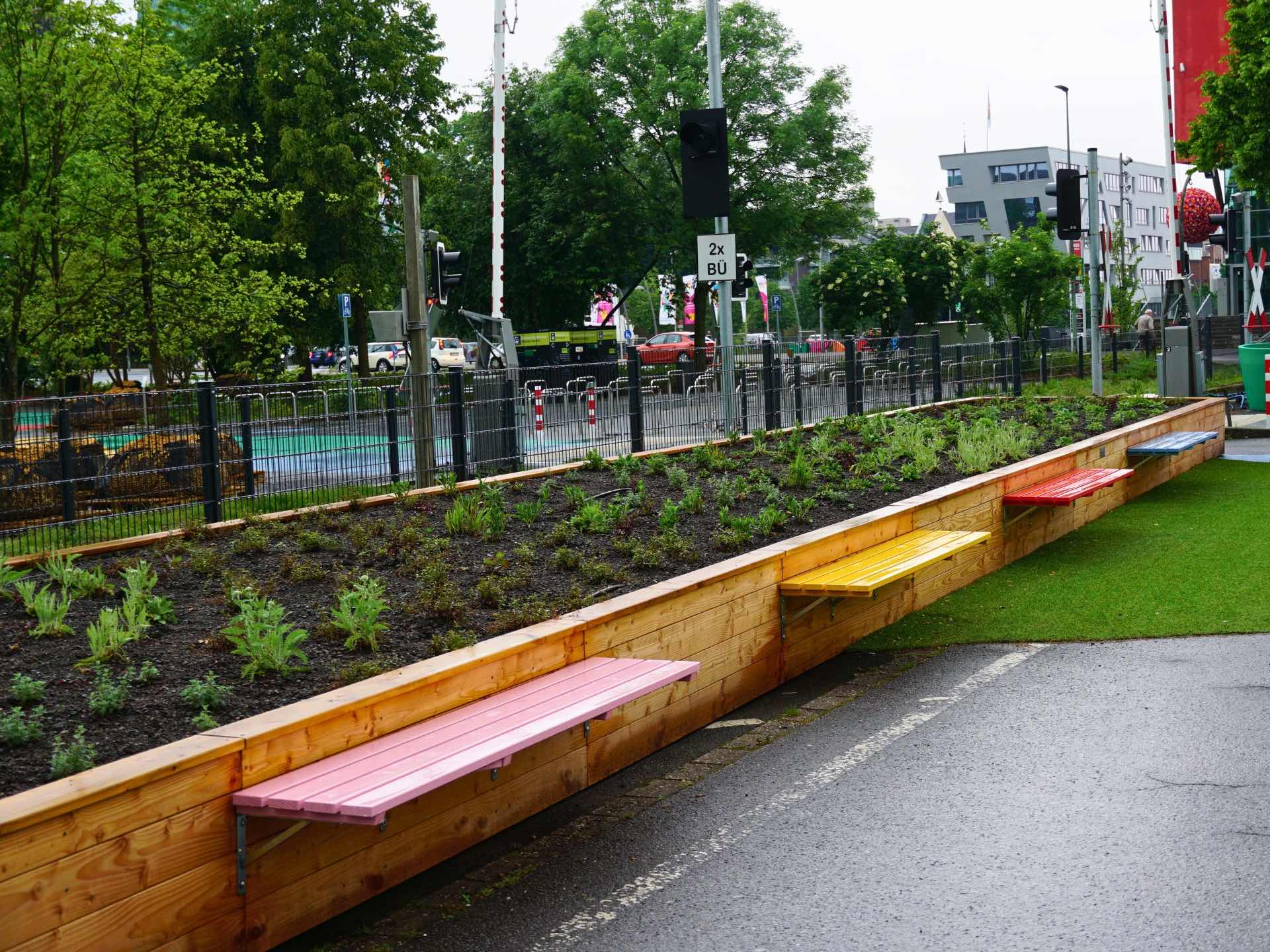 Polygonales Hochbeet aus naturbelassener Douglasie mit integrierten Sitzbänken in Hellrosa, Signalgelb, Reinrot und Pastellblau – Referenzprojekt Landesgartenschau Neuss 2026