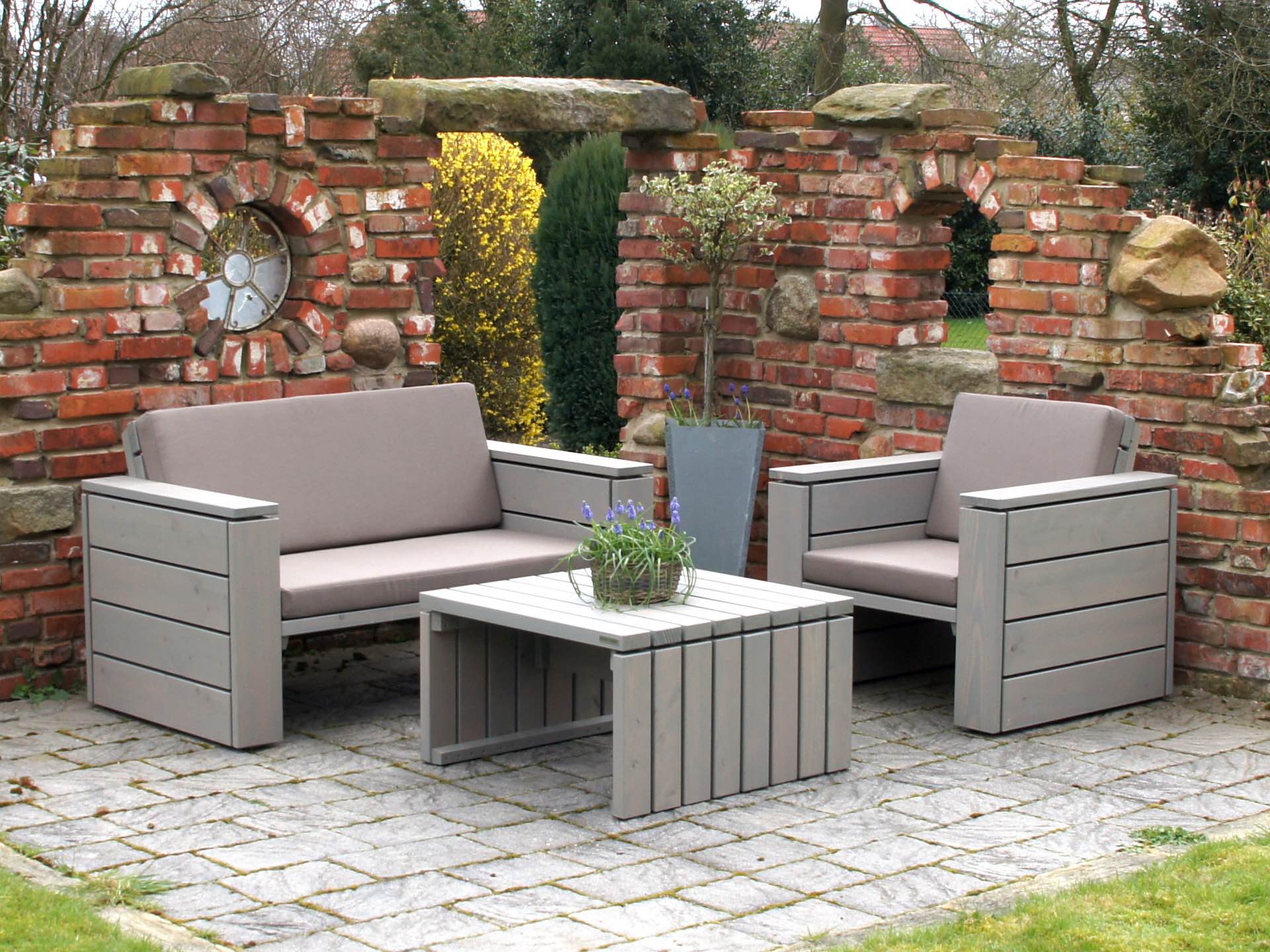 Loungemöbel-Set 1 grau geölt mit Sofa, Tisch und Sessel, sandfarbene Polster, auf Natursteinen vor Klinkermauer