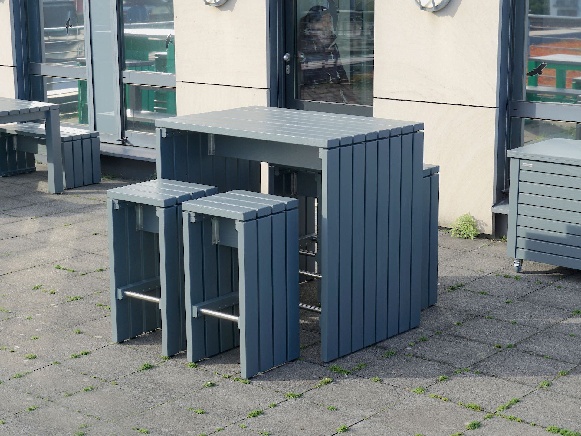 Barmöbel-Set aus Holz in Grau lackiert mit Tisch und vier Barhockern vor Fassade eines Bürogebäudes