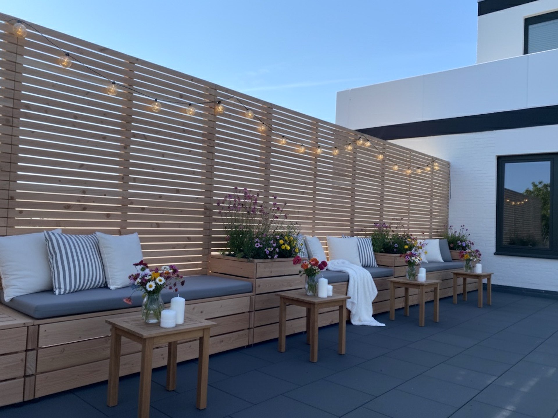 Moderner Sichtschutz mit Pflanzkästen & Sitzbänken für die Terrasse Holzsichtschutz im Garten mit Pflanzkästen, Sitzbänken, dekorativer Lampion-Lichterkette, bunten Blumen, Kerzen und bequemen Polstern.