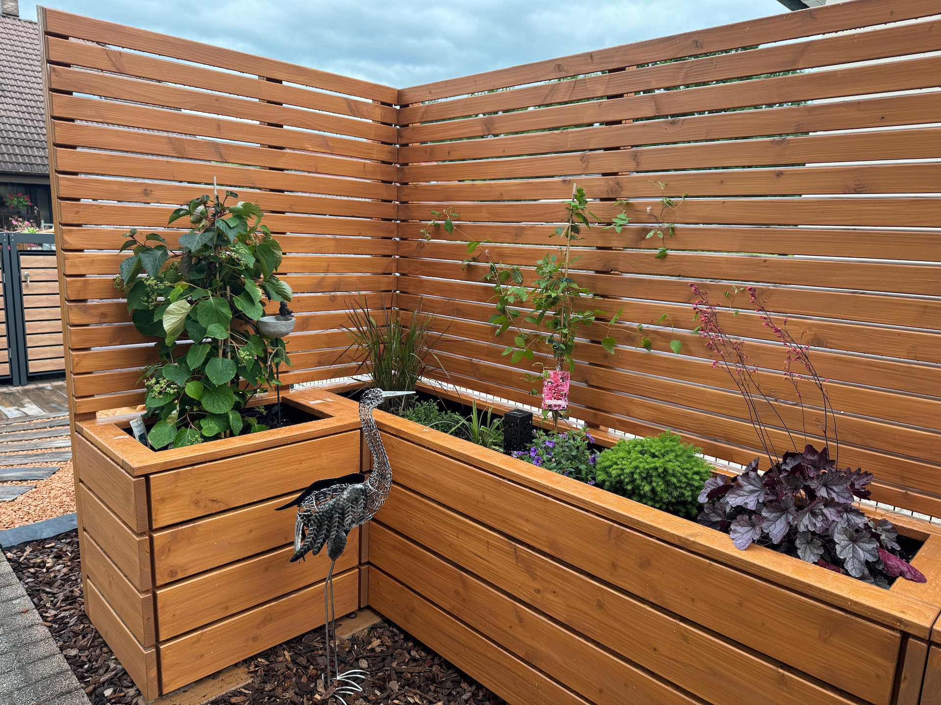 Gestaltungsbeispiel mit bepflanzter Sichtschutzecke und Rindenmulchfläche Eingebundene Sichtschutz-Ecke mit harmonisch bepflanzten Kästen und strukturiertem Gartenbereich