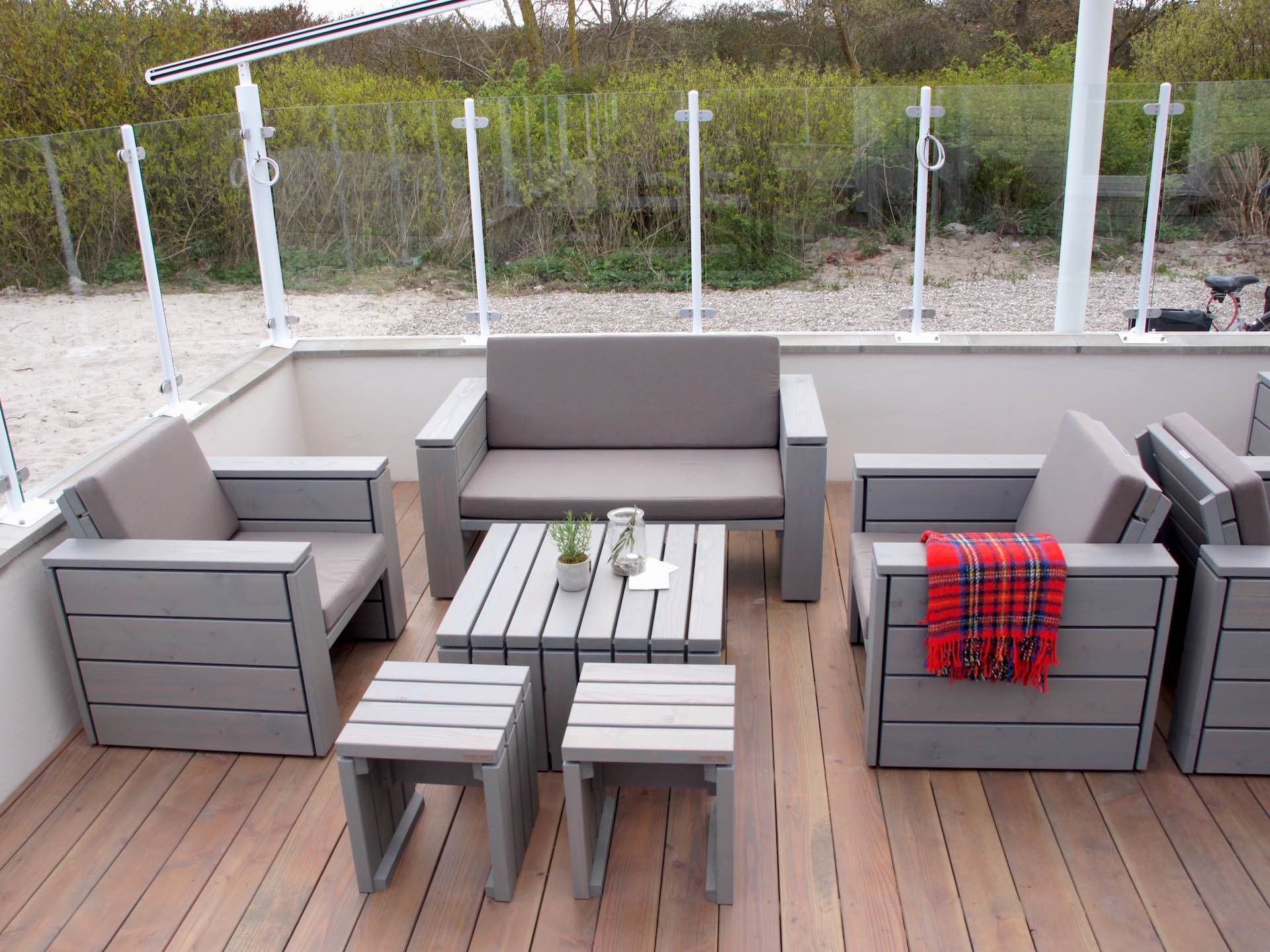 Loungemöbel auf Café-Terrasse, grau geölt, mit Polstern in Sand, bestehend aus 2er Sofa, zwei Sesseln, zwei Hockern und Tisch