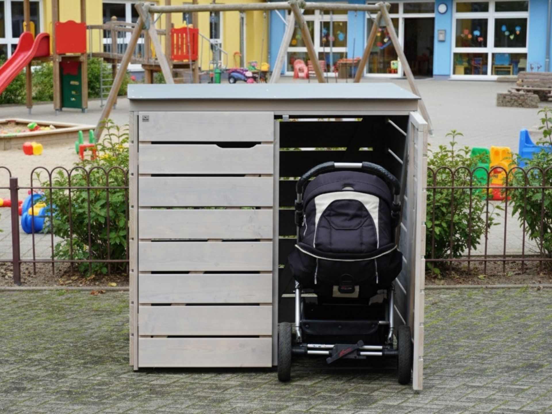 Große Kinderwagenbox aus grau geöltem Holz mit HPL-Dach, eine Tür geöffnet, Kinderwagen steht davor – wetterfeste Lösung für Kitas.
