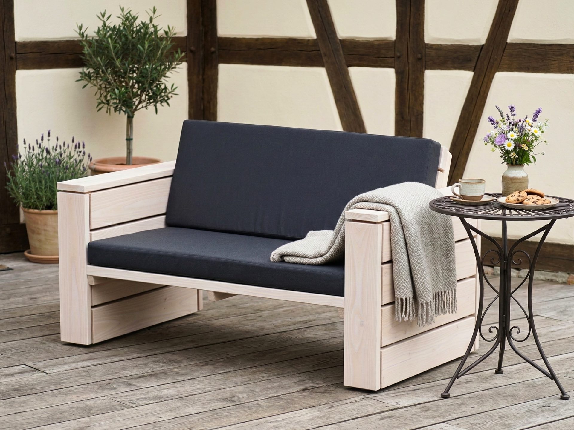 Weiß geöltes Lounge Sofa 2-Sitzer auf Terrasse neben Metalltisch mit modernem Materialmix aus Holz, Stoff und Metall.