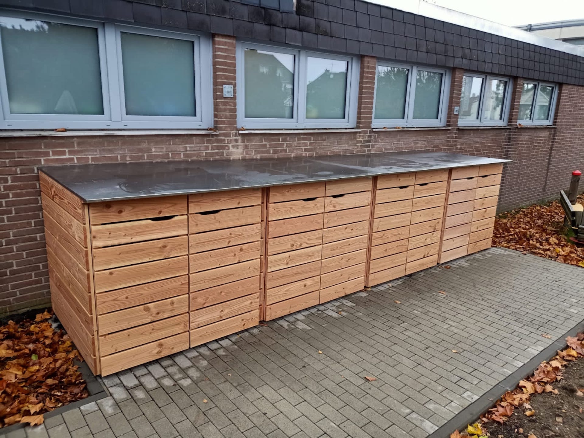 Kinderwagenbox mit 8 Stellplätzen aus Naturholz und Edelstahl-Dachverkleidung, auf Pflasterfläche vor einer Kita