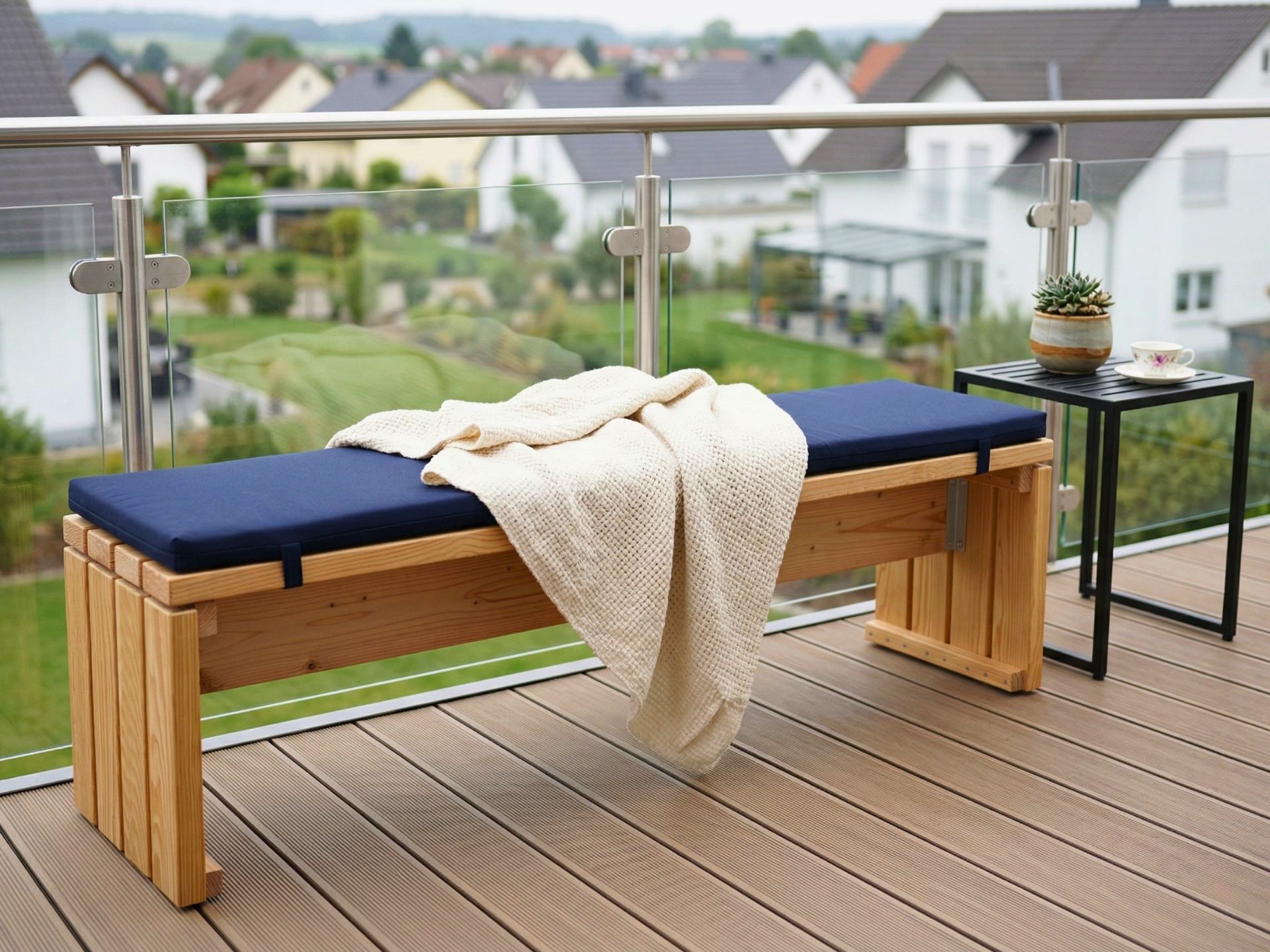 Gartenbank aus Douglasie mit blauem Polster auf Balkon, daneben Metalltisch mit Blumentopf und Kaffeetasse.